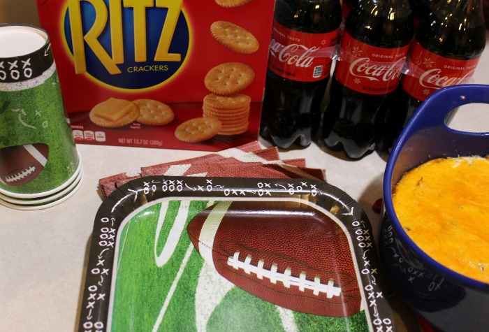 Creamy Cheddar Bacon Dip next to football paper plates, a box of ritz, and bottles of coca cola