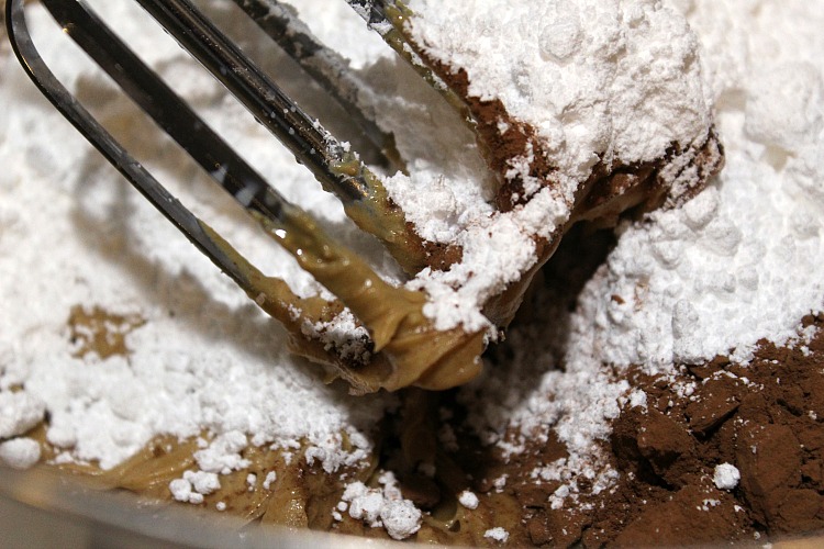 Peanut butter, milk, and butter mixed with an electric hand mixer with baking cocoa and powdered sugar in a mixing bowl
