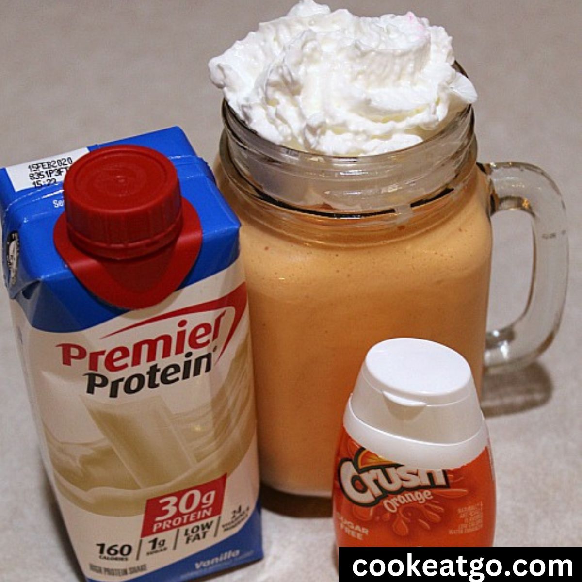 Mason Jar Mug with orange creamsicle protein shaek topped with whip topping next to a carton of vanilla premier protein and orange crush drops 