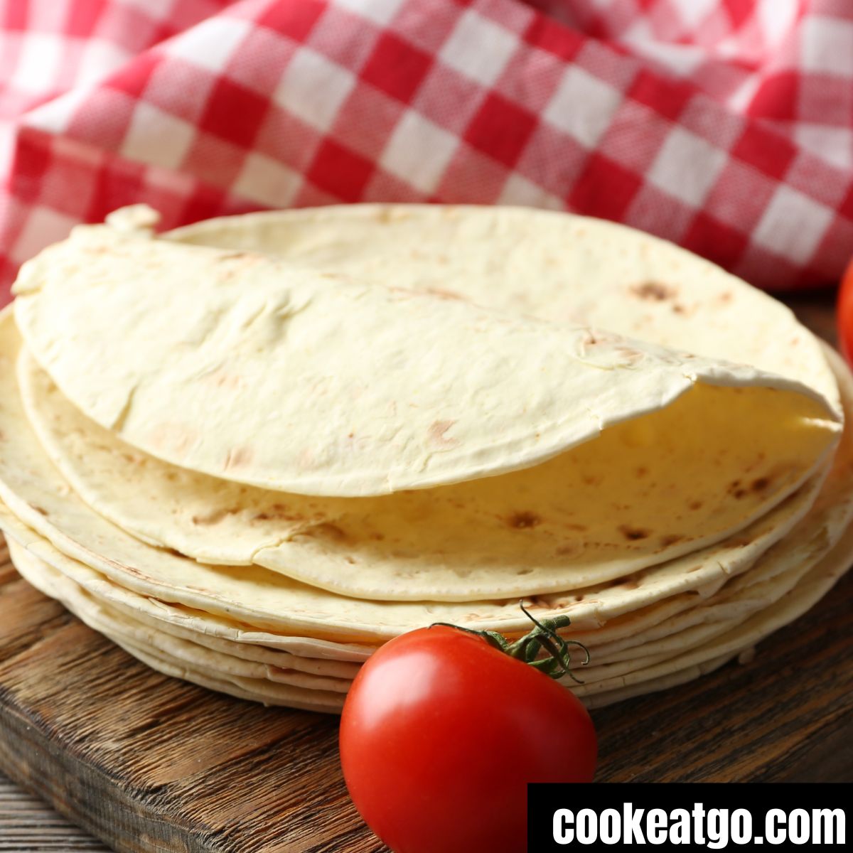 Stack of tortillas next to a tomato on a board with a red white checkered napking