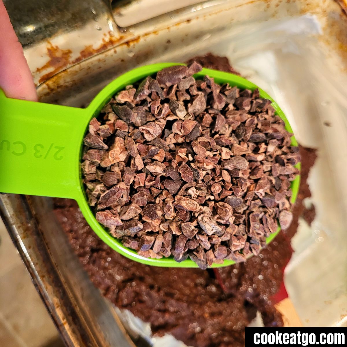 Measuring cup of viva naturals cacao nibs over black bean brownie in 8x8 pyrex pan